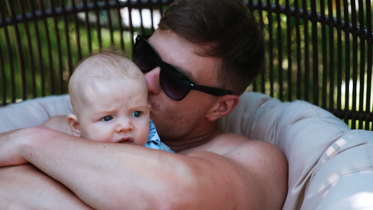 Father embracing a cute baby boy in his arms. Dad in sunglasses kisses his lovely infant son sitting outdoors. Close up.