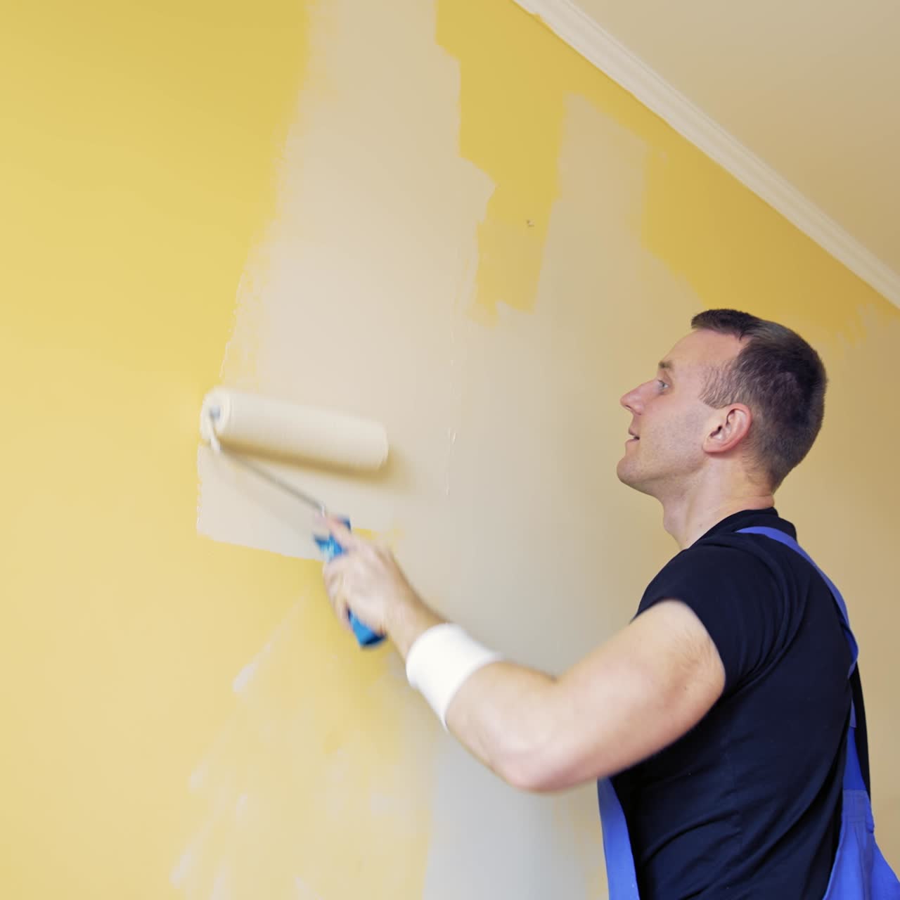 Room renovations at house. Paint being applied on a wall by a professional worker. Painter man painting the wall at home with paint roller and creamy color paint. Side view