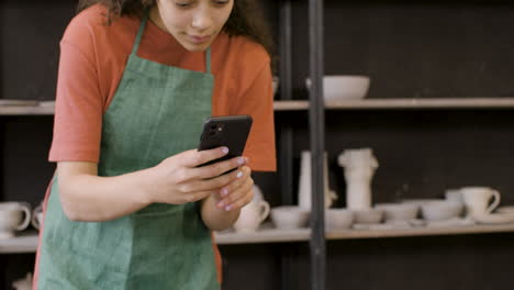 mujer sonriente en delantal usando teléfono y tomando fotos de piezas de cerámica hechas a mano en la mesa en la tienda de cerámica