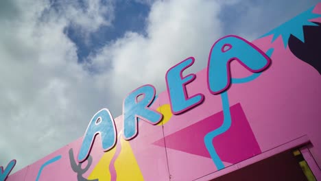 funky-colourful-sign-text-written-area-in-blue-for-families-kids-on-purple-yellow-background-at-festival-designated-area-with-partly-clouded-sky-in-the-background-orbital-camera-movement