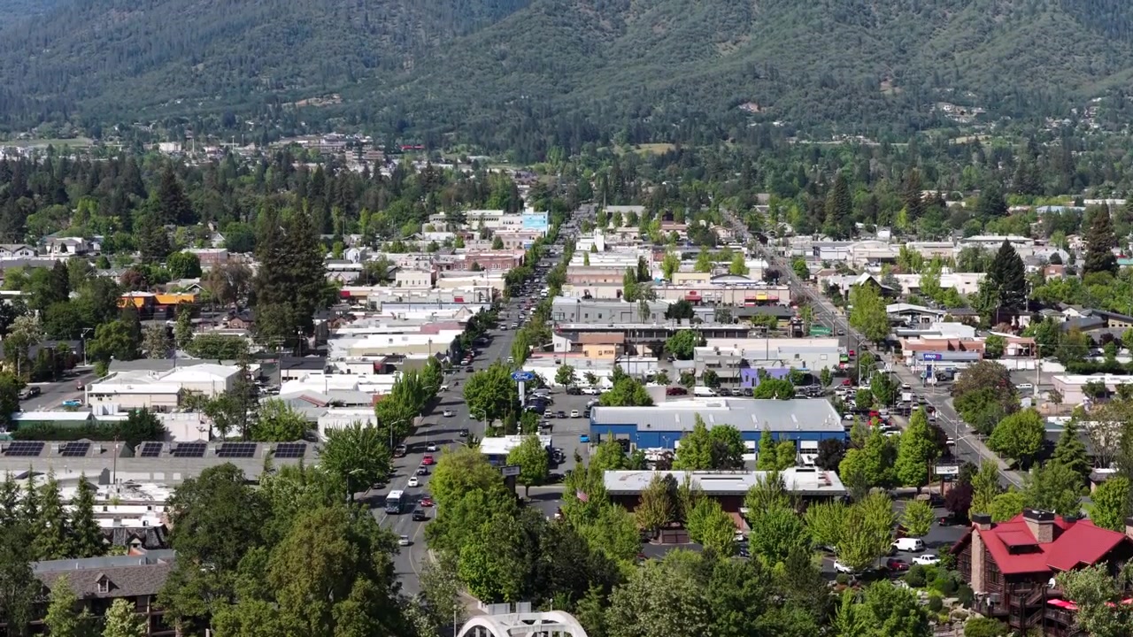 Centro di Grants Pass, Oregon, con storico ponte ad arco sul fiume Rogue, folta chioma verde di alberi, quartiere commerciale a bassa densità, quartieri residenziali e colline boscose sotto un cielo soleggiato. Salita aerea.