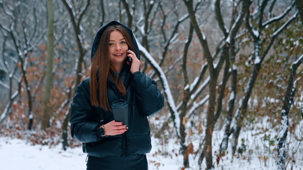 A walk in the winter park with a cup of hot coffee. Young dark-haired lady speaks on the phone having a stroll outdoors.