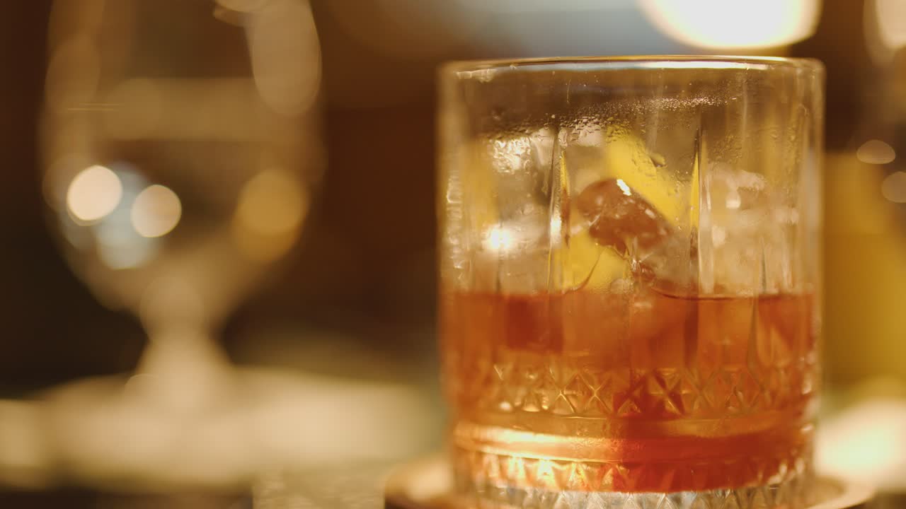 A hand picks up a classic Negroni cocktail from a bar table in warm, low lighting, with a blurred glass in the background