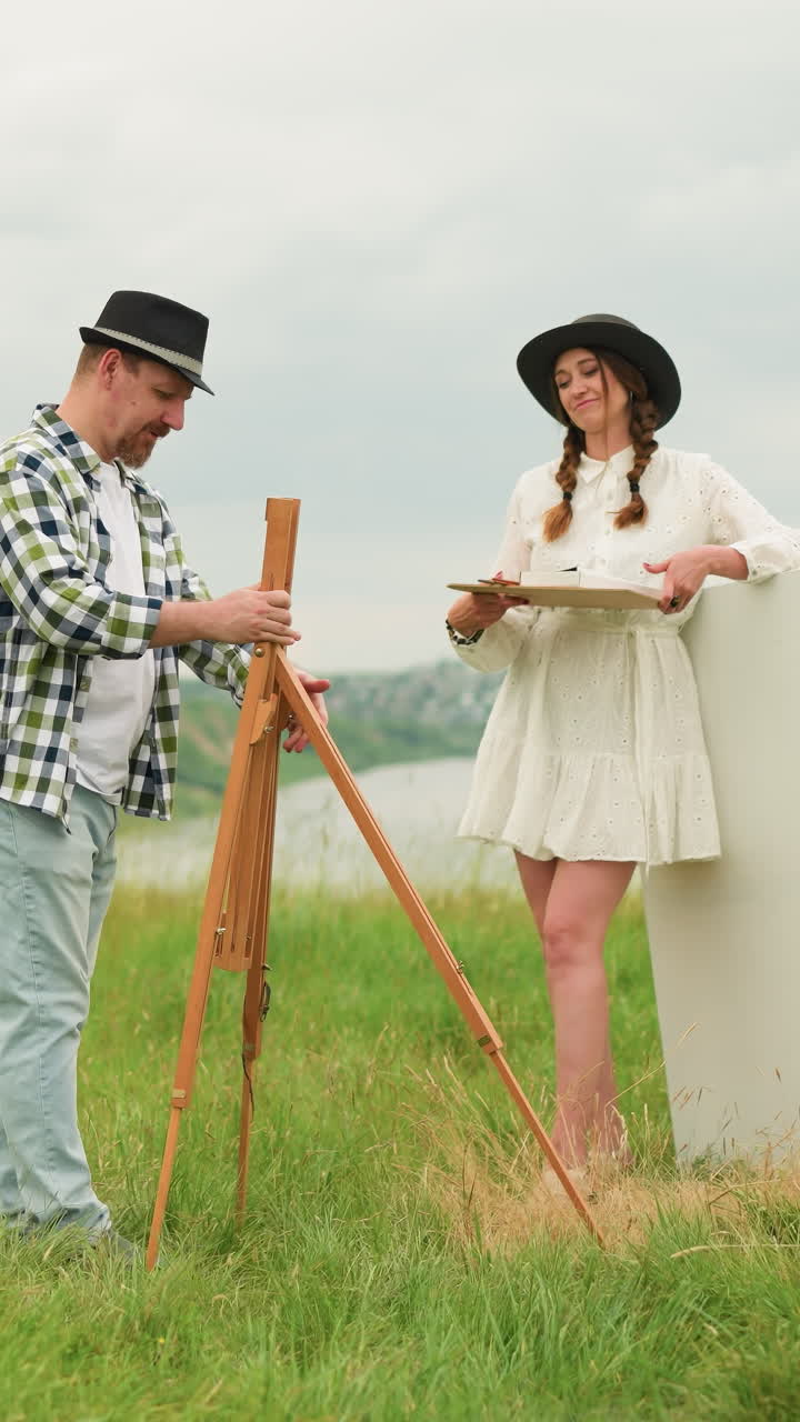 una pareja se prepara para pintar al aire libre en un campo verde exuberante con un. el hombre, con un sombrero negro y una camisa a cuadros, establece un caballete mientras que la mujer, con un vestido blanco y un sombreiro negro, sostiene una paleta de pintura