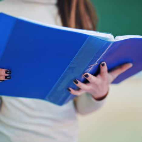 blue folder in woman's hands in class. woman turns pages with printed papers in files on educational center background. close-up.