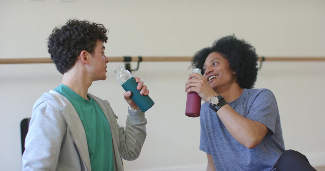 African American trainer with teen male trainee smiling, drinking bottles after coach in studio