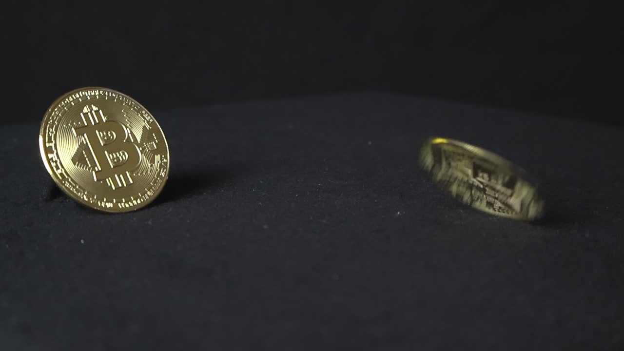 dos monedas de criptomoneda de color amarillo dorado reflectante en un fondo oscuro, mientras que una está inmóvil en exhibición y la otra está girando a cámara lenta creando sombras y parpadeos de luz.