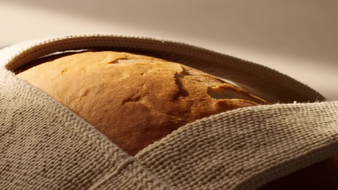 Close-up of a crusty artisan bread loaf with a woven fabric strap across the foreground, shown in warm soft light against a blurred neutral background