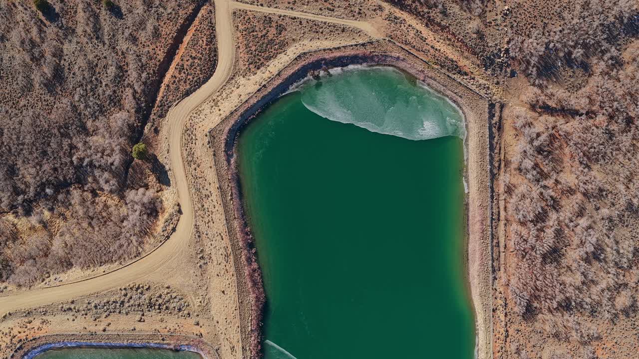 A high-angle drone shot looks directly down at an emerald green reservoir with thin, white ice sheets. The 4K view captures the stark contrast between the deep water and the arid desert shoreline