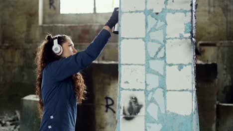 female graffiti painter is creating abstract image on pillar in old damaged warehouse and listening to music with headphones. modern art and creative people concept.