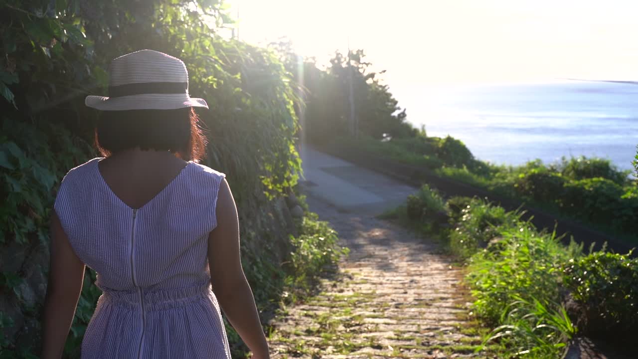 chica en verano fuera con sombrero caminando por el camino hacia el océano con sol quema lenta