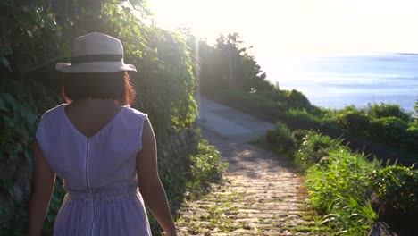 girl in summery outwith wearing hat walking down path towards ocean with sun flaring slow motion