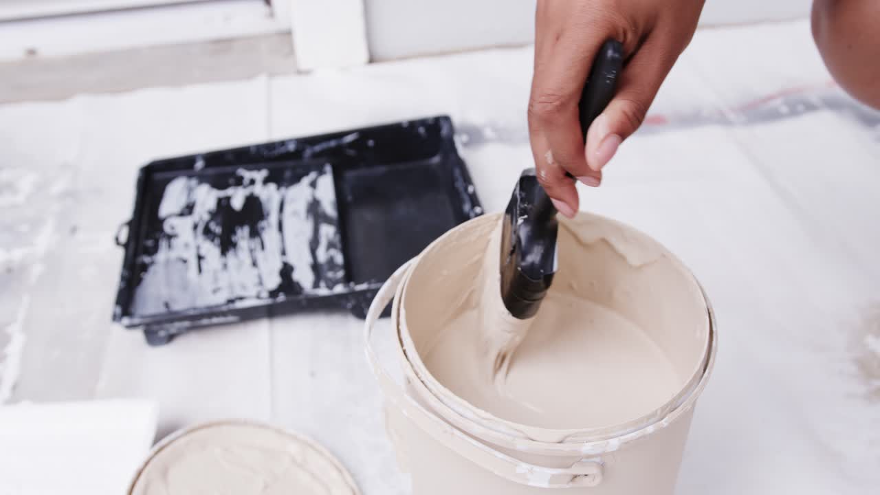 African American woman dipping tool, stirring beige paint in bucket on drop cloth preparing coat