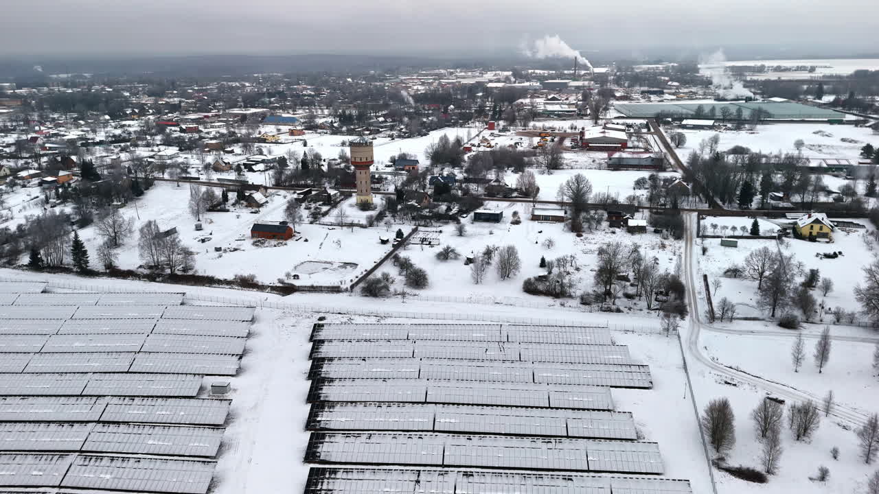 High drone shot with camera panning from left to right over snow-covered solar panels, a water tower, scattered houses, roads, and distant industrial smoke in a winter landscape