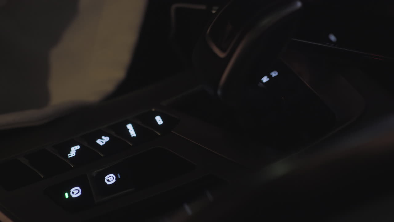 Close up of car gear selector and illuminated dashboard buttons including electronic parking brake, auto hold, drive mode and sport mode with blurred fabric detail visible in soft ambient lighting
