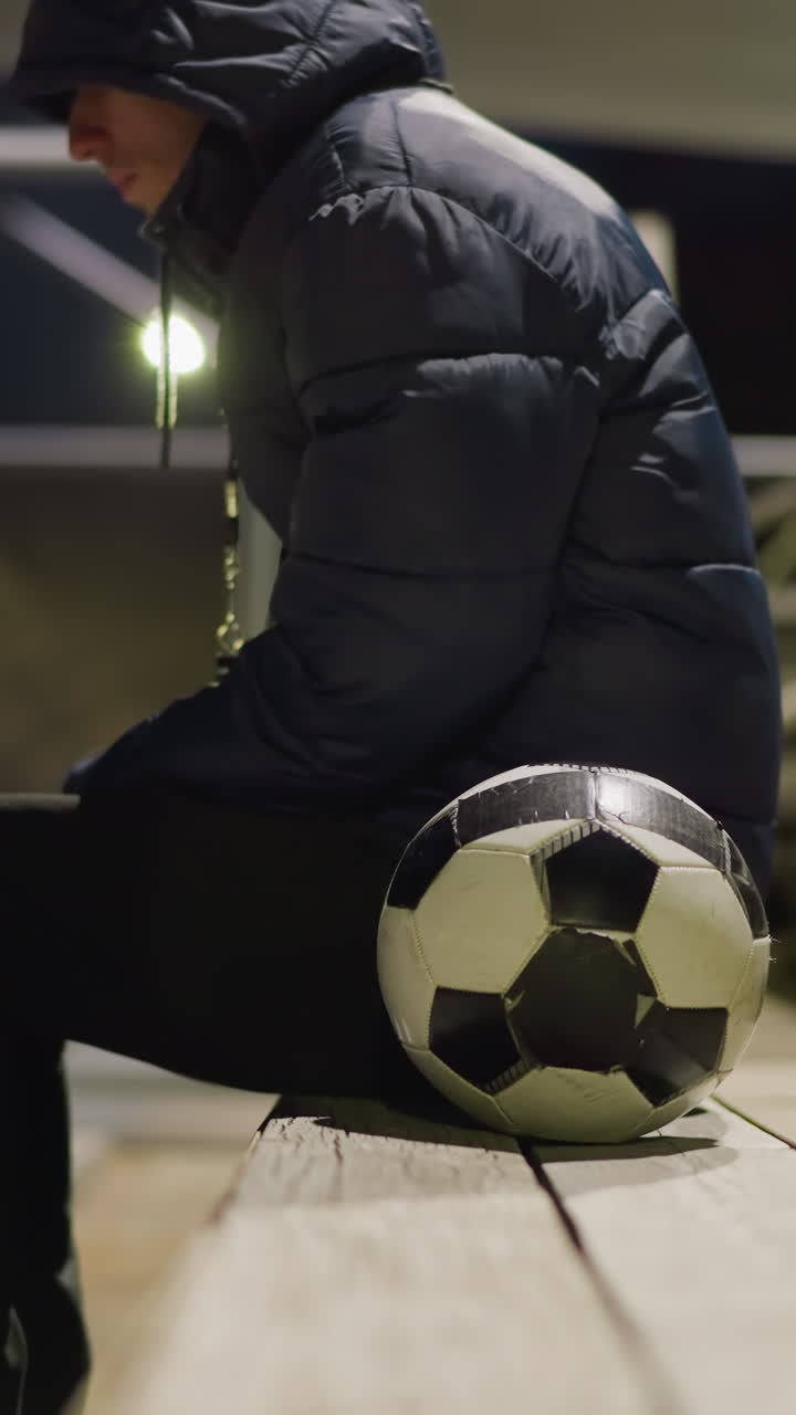 близкий боковой вид человека в черном наряде и куртке, сидящего в одиночестве на тихом, тускло освещенном стадионе, он сидит с руками между ногами, с футбольным мячом рядом с ним