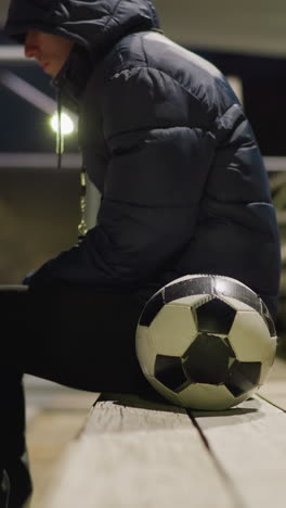 vista lateral de primer plano de un hombre con un traje negro y una chaqueta sentado solo en un estadio tranquilo y débilmente iluminado, está sentado con las manos entre las piernas, con una pelota de fútbol a su lado