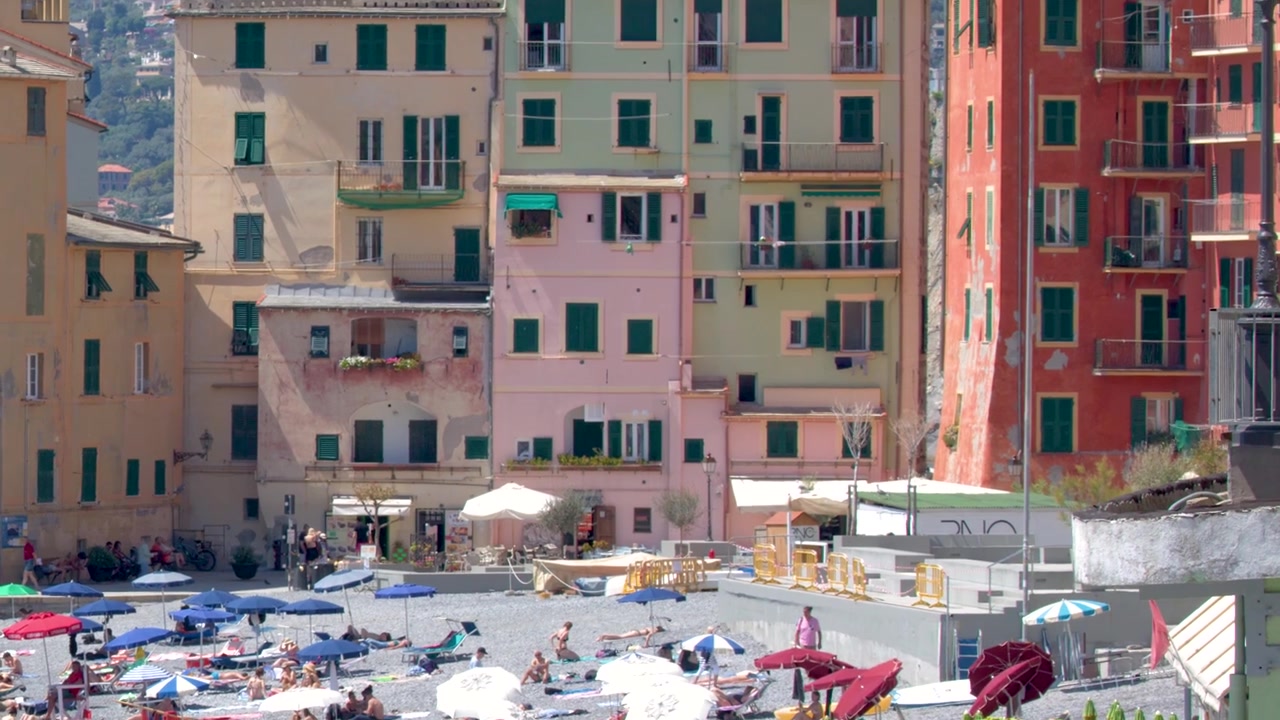 Una lenta inclinazione verso l'alto rivela una vivace architettura costiera che si affaccia su una affollata spiaggia di ciottoli mediterranea.