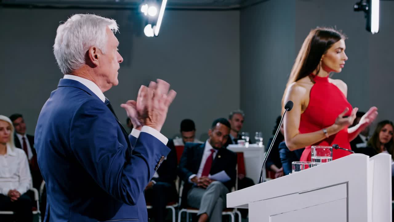 Un fotogramma video di una presentazione aziendale. L'angolo laterale riprende un uomo in giacca e una donna in rosso