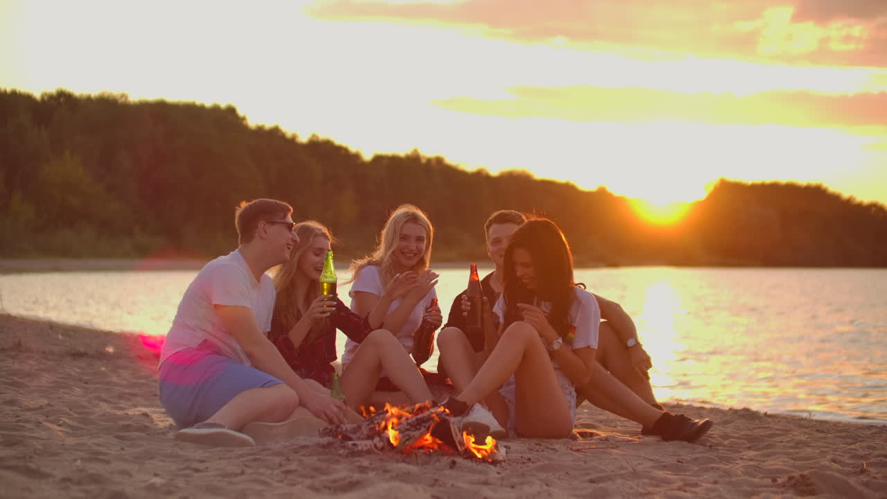 Five young people are sitting around bonfire on the sand beach. They are talking to each other and drinking beer at sunset and enjoying the summer evening on the lake coast.