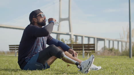 tiro largo de un deportista cansado con una pierna artificial bebiendo agua en el césped del parque y relajándose después de un intenso entrenamiento cardiovascular por la mañana 1