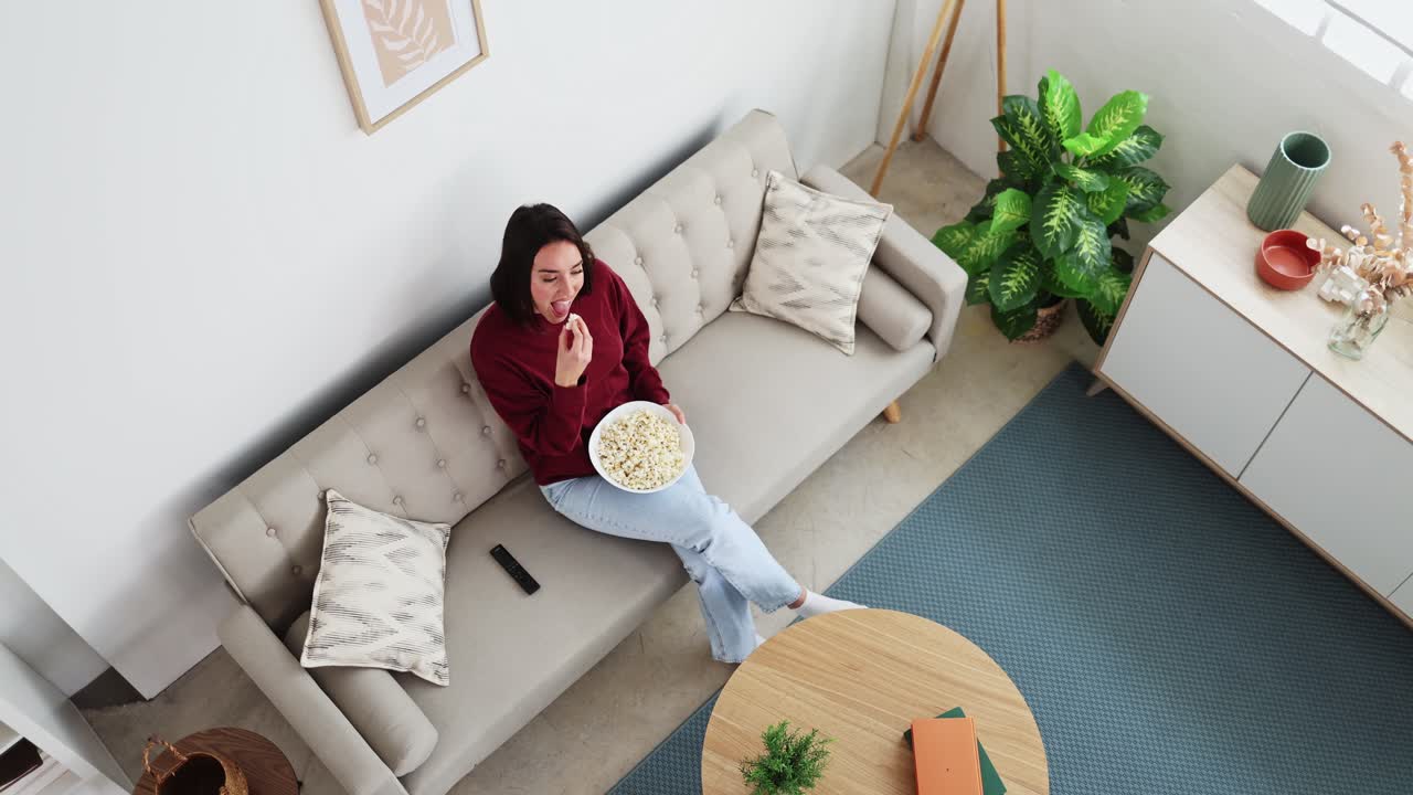 Ung kvinde slapper af på sofaen, spiser popcorn og ser tv
