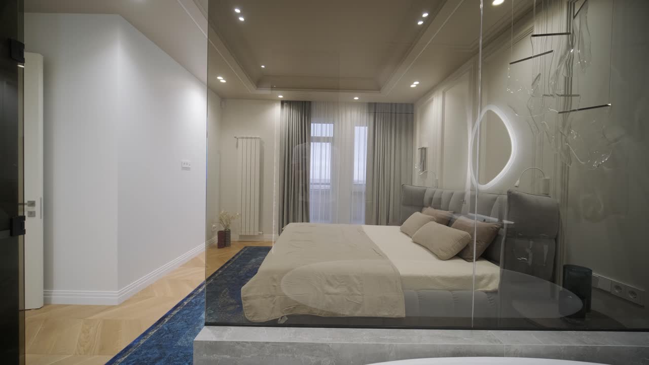 A luxurious bedroom with a stylish tufted headboard and a striking blue rug seamlessly transitions into a sleek bathroom with modern fixtures and a unique oval mirror, capturing a design that melds comfort with elegance.