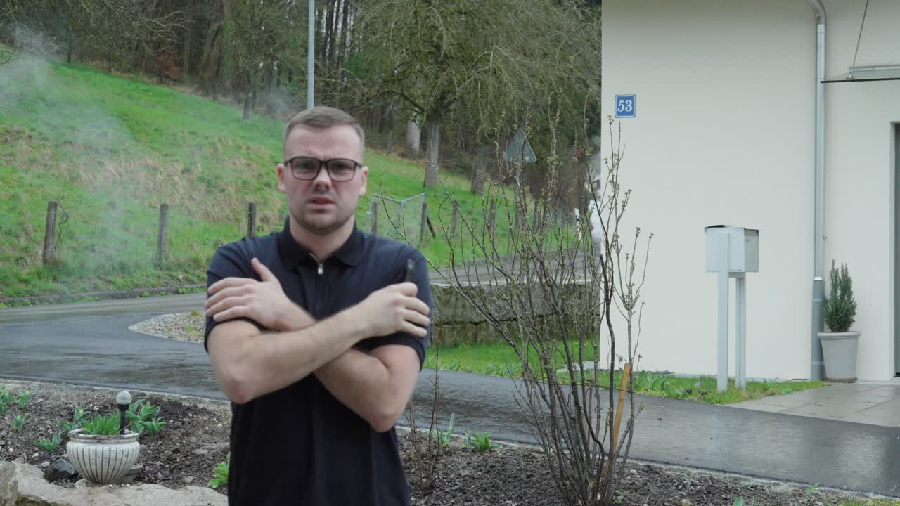 Ung mand står udendørs i koldt vejr, mens han ryger og fryser, hvilket viser vinterubehag, hverdagsvane og realistisk hverdagsliv i udendørs omgivelser