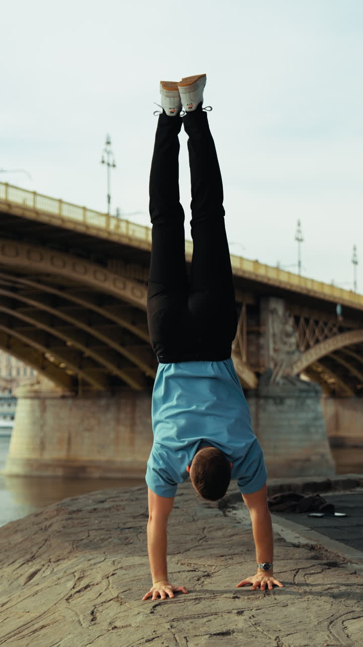 Giovane uomo che esegue un esercizio di verticale lungo il fiume Danubio con il Ponte Margherita sullo sfondo, Budapest. Fitness all'aperto, allenamento dell'equilibrio e stile di vita attivo.