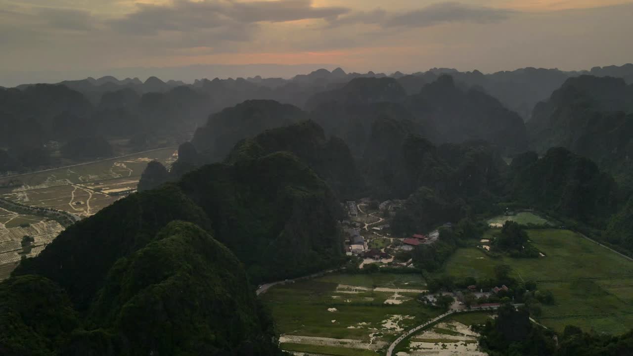 Village tucked among karst hills and patchwork rice fields in Ninh Binh glows toward dusk as clouds drift and paddies mirror the sky over the valley, forward moving drone timelapse