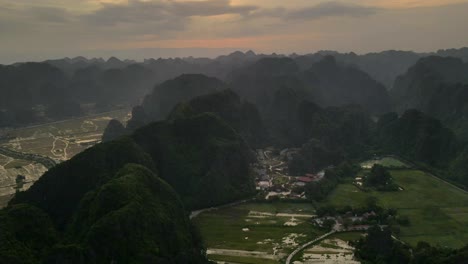 village tucked among karst hills and patchwork rice fields in ninh binh glows toward dusk as clouds drift and paddies mirror the sky over the valley, forward moving drone timelapse