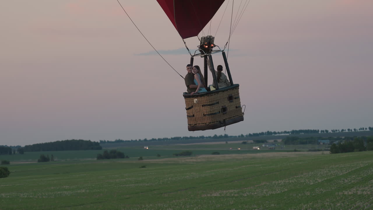 passengers ride hot air balloon basket over expansive green field at soft dusk flame bursts upward illuminating scene farm land below distant car headlights glimmer
