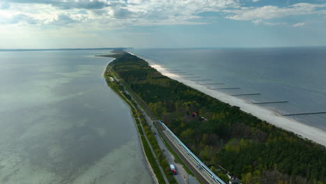 aerial view capturing the unique geography of kuźnica on the hel peninsula, showing the narrow strip of land with a railway line running alongside lush greenery, bordered by the baltic sea and bay