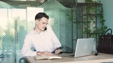 portrait of a handsome, young, stylish man in business attire sitting at a desk with a laptop in a modern office, talking on the phone and taking notes in a notebook, busy and focused on work