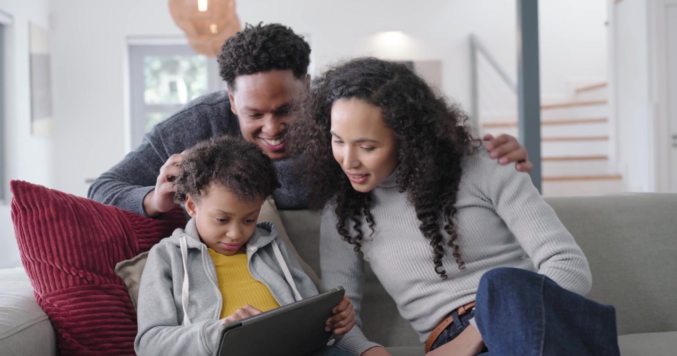Famiglia afroamericana che reagisce al bambino che tocca il tablet, con i genitori che si sporgono dal divano guidando le sue mani