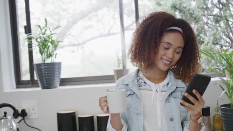 happy biracial woman using smartphone and drinking coffee in kitchen, slow motion