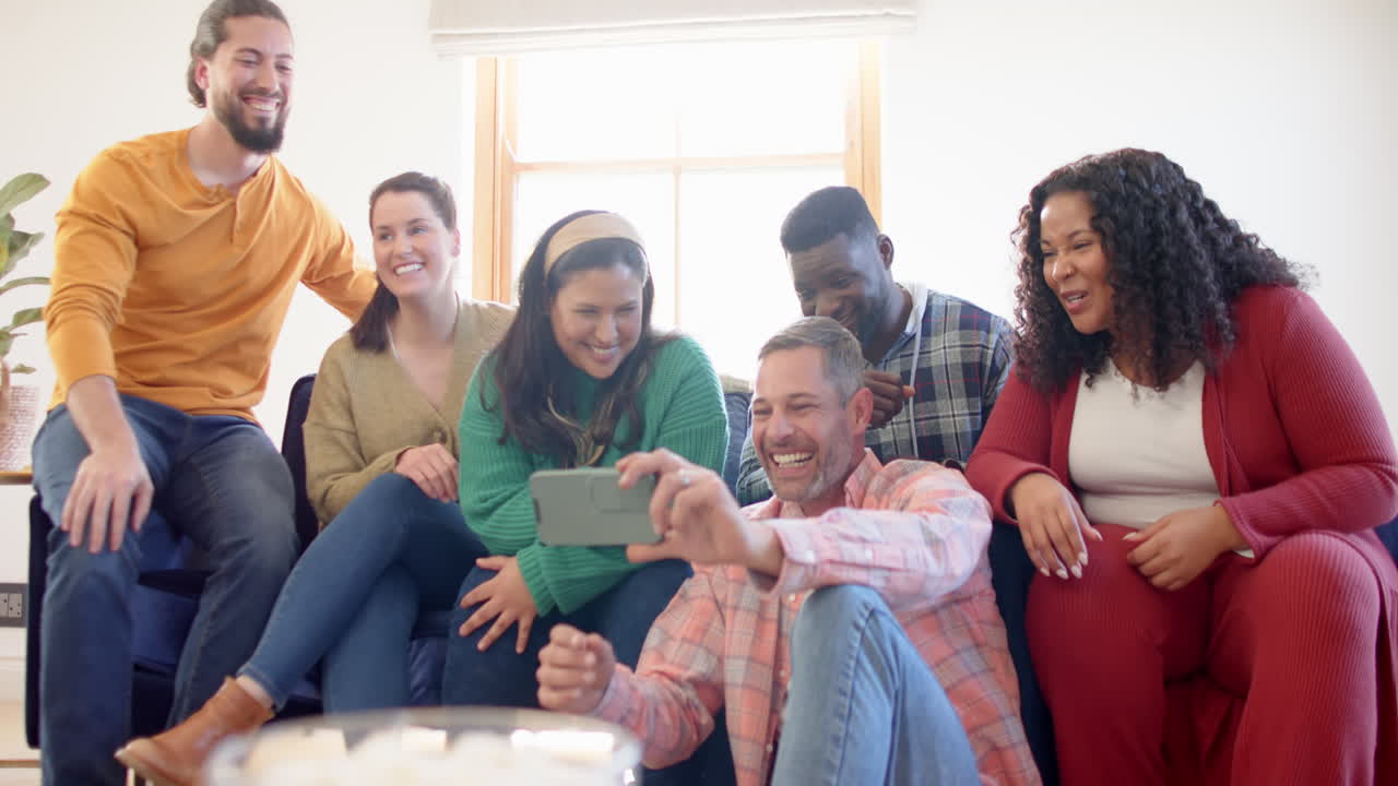 Felices amigos y amigas diversos relajándose en casa juntos hablando y mirando el teléfono inteligente