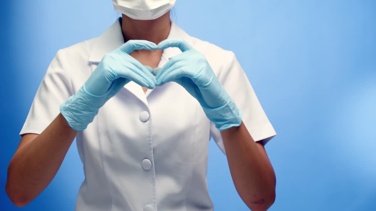 una enfermera con mascarilla y uniforme blanco hace un signo cardíaco con las manos - fondo azul con espacio para copiar