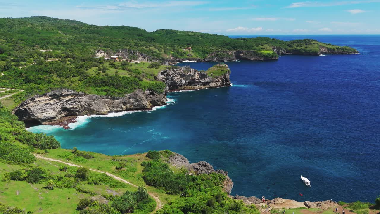 Forward-rising drone shot reveals Nusa Penida’s rugged limestone coastline at Manta Bay near Broken Beach, with vivid turquoise water, reef edges and lush green headlands