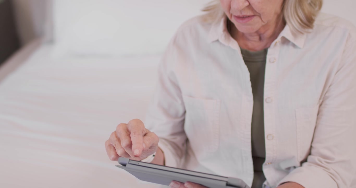 Senior woman noticing tablet on lap, positioning and tapping, swiping screen while reading on bed