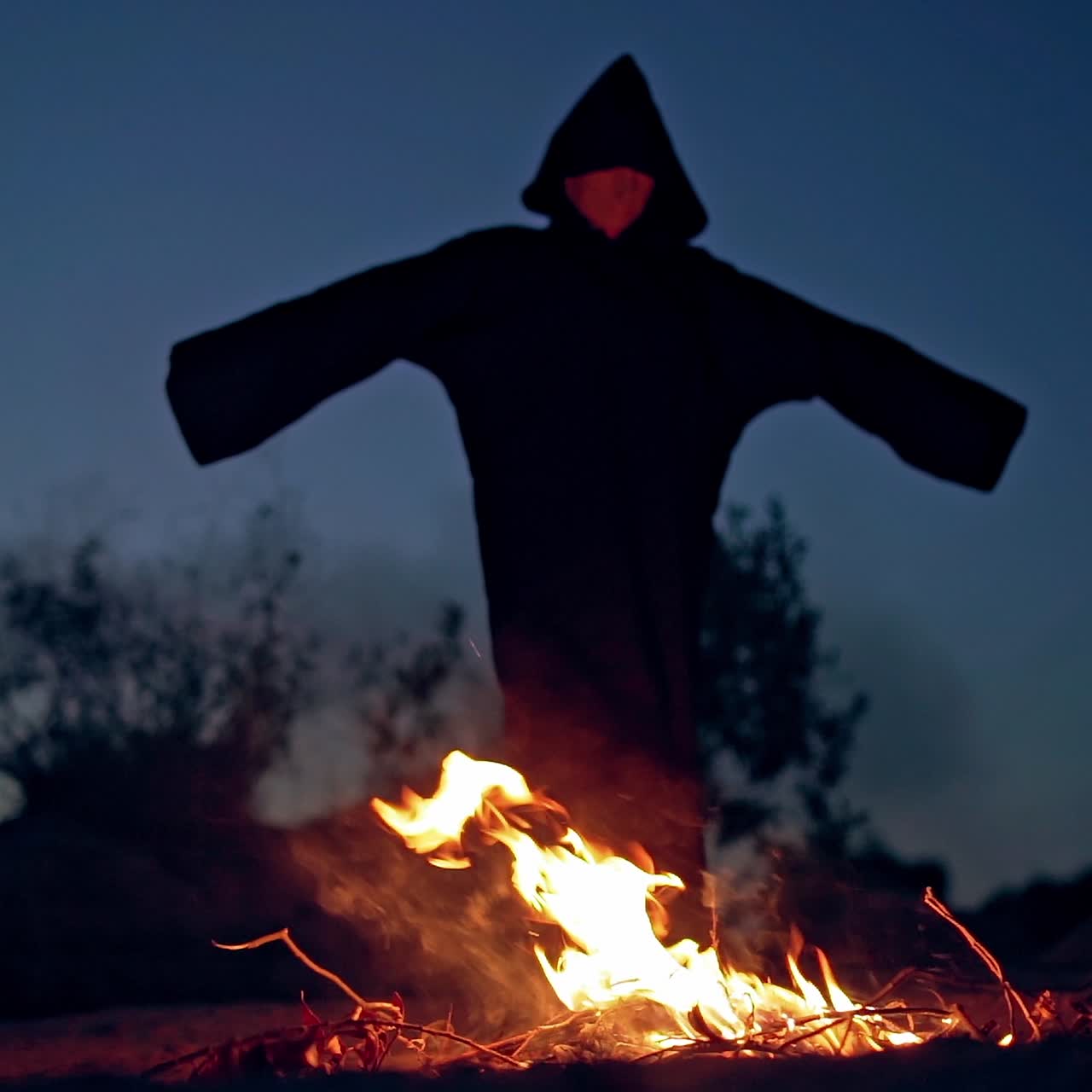 Scary witch stands by the fire at night. Black figure in cloak with hood walking around the fire with outstretched arms. Halloween outdoors.