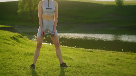 close-up: a female sportswoman squats with dumbbells against the backdrop of a lake in the park in sportswear. morning training on the street.