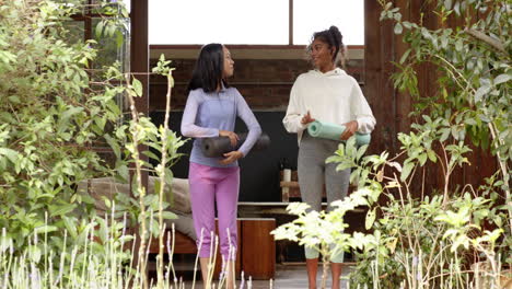 two multiracial female friends with yoga mats smiling and chatting in garden setting