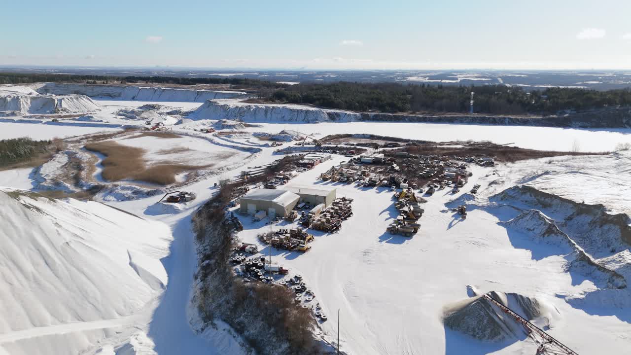 заснеженный карьер с оборудованием, кучами для хранения и отдаленным лесистым горизонтом