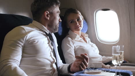 vista lateral de una alegre mujer de negocios y un hombre de negocios barbudo charlando y tomando copas de champán mientras viajaba en clase ejecutiva en avión