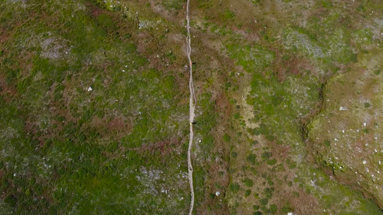 Top down aerial view of a lonely mud hiking road or pathway going straight across a grassy green and brown Sweden landscape into the distance during a cloudy day. The ground has big rocks and moss.