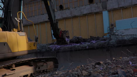 A-large-yellow-Volvo-excavator-is-digging-and-pulling-from-the-ground-dirt-hole-at-a-demolition-construction-building-work-site-in-the-sun-tearing-the-concrete-wood-plywood-wall-in-front-slow-motion