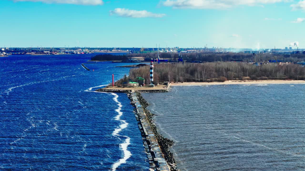 A black and white striped lighthouse stands on a narrow peninsula separating a blue sea from a brown river estuary. The clear sky and distant cityscape complete the scenic view.