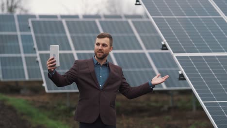 Un-Hombre-Sólido-Vestido-De-Negocios-Está-Hablando-Con-Una-Tableta-En-El-Fondo-De-Una-Planta-De-Energía-Hecha-De-Paneles-Solares