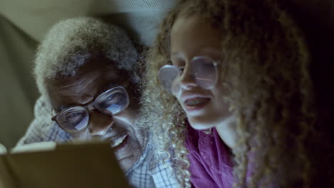 Cute-girl-and-her-grandpa-reading-book,-hiding-under-blanket